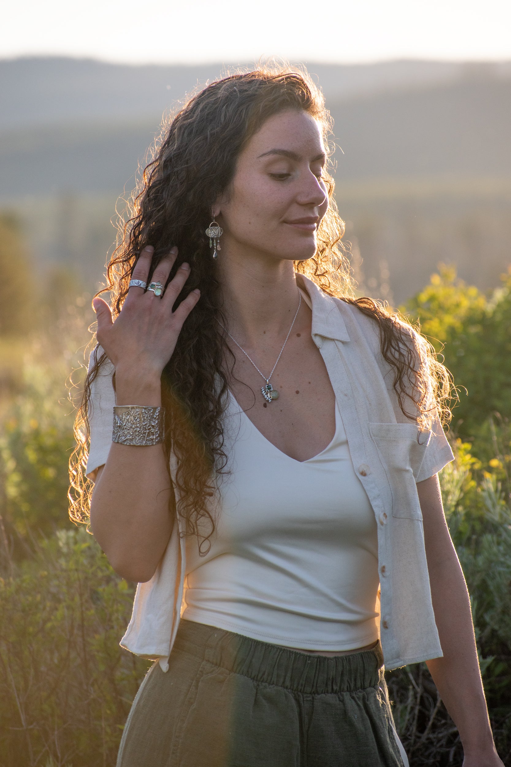 A woman outdoors at sunset wearing a white tank top, an open light-colored shirt, olive green pants, and silver jewelry including a necklace, bracelet, and rings.
