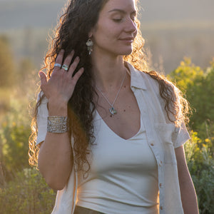 A woman outdoors at sunset wearing a white tank top, an open light-colored shirt, olive green pants, and silver jewelry including a necklace, bracelet, and rings.