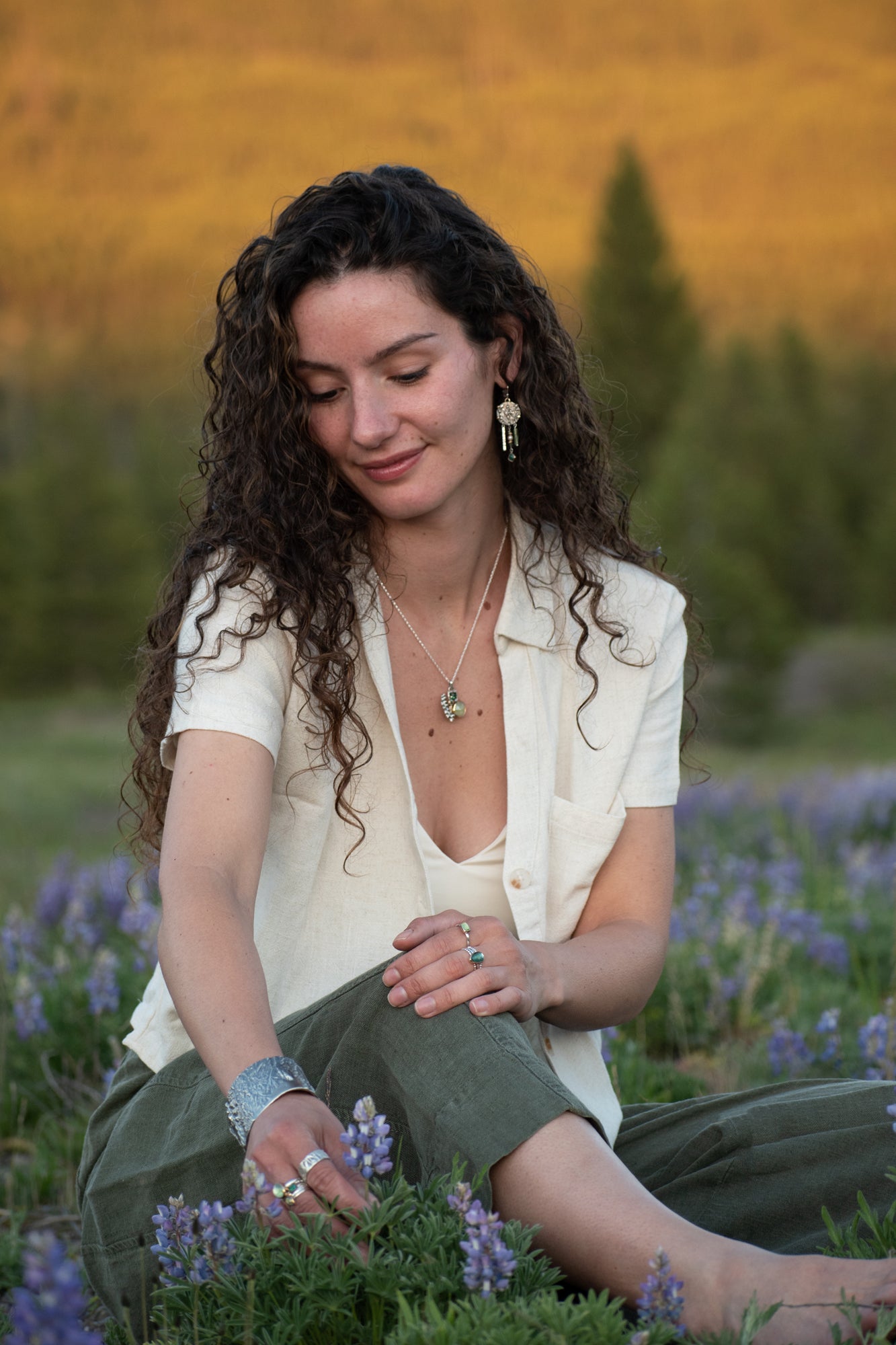A woman with long curly dark hair sits in a field of purple flowers, wearing wild meadow, nature inspired jewelry. The background features a soft-focus landscape of golden hills and green trees.