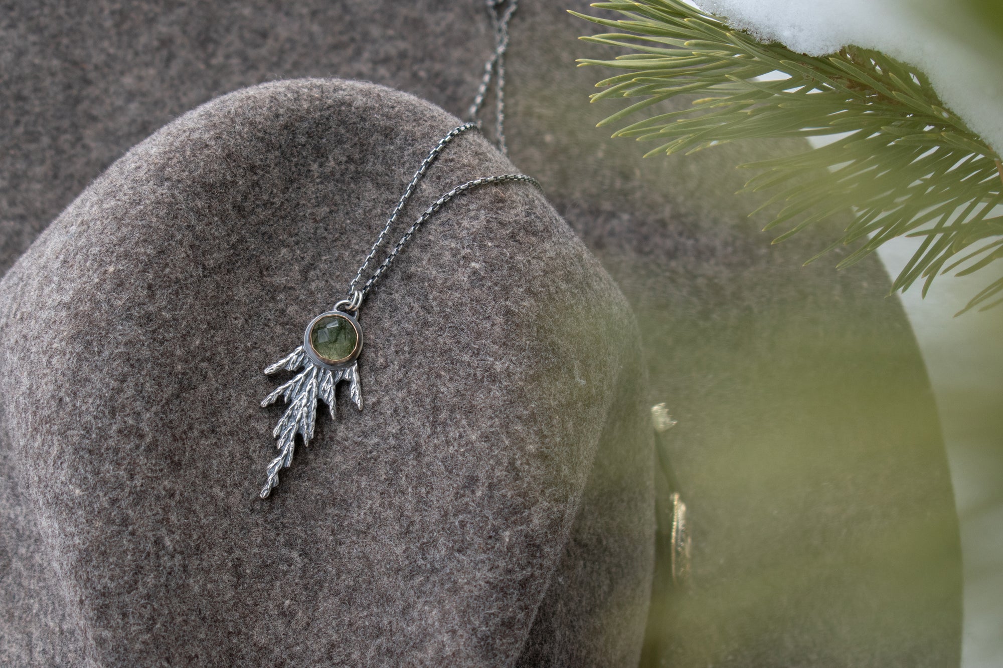 Handmade sterling silver cedar necklace with green stone set in 14k gold draped on women's hat with evergreen in foreground