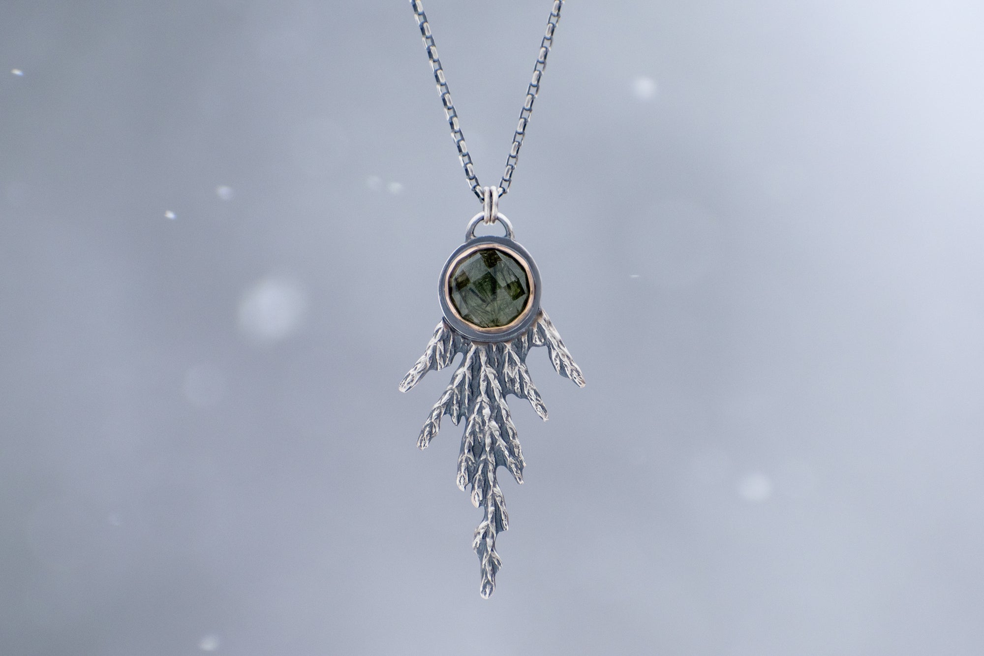 Handmade sterling silver cedar necklace with green rutile set in 14k gold hanging in snow